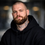 Bearded man with neck tattoos in a black hoodie holding a magnetic phone mount, facing the camera.